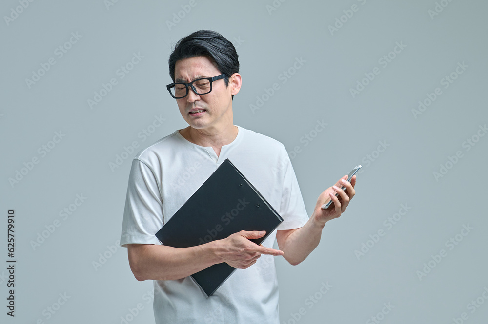 A middle-aged office worker wearing casual clothes and glasses is holding a smartphone and files and making various emotional expressions. A middle-aged office worker wearing casual clothes and glasses is holding a smartphone and files and making various emotional expressions.