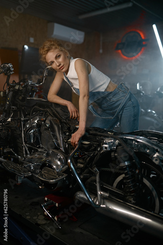 Beautiful woman mechanic posing for camera nearby motorcycle at workshop