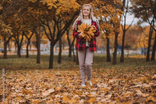 middle aged woman plays with autumn leaves. Mature blonde woman in a plaid shirt with a bouquet of yellow leaves in the park in autumn enjoys life  walks and throws leaves