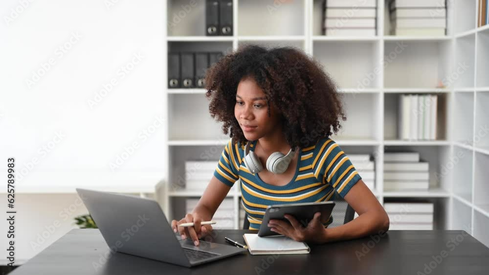 Attractive happy young African student studying at the college library, sitting at the desk, using a laptop computer, tablet and headphones having a video chat. in room.. Attractive happy young African student studying at the college library, sitting at the desk, using a laptop computer, tablet and headphones having a video chat. in room..