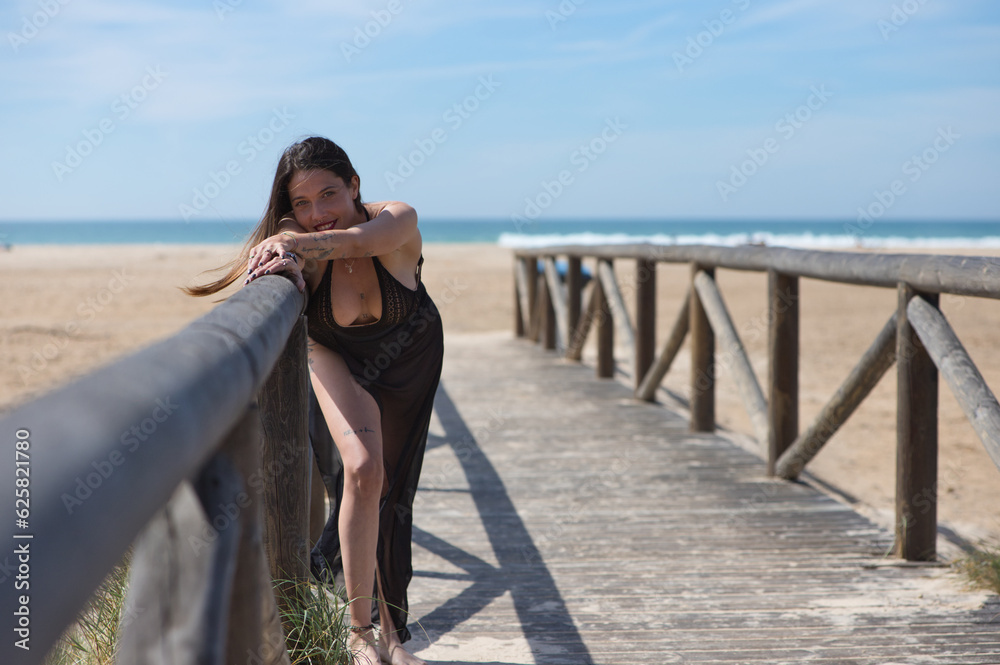 Young, beautiful woman with long brown hair, wearing a swimming costume and a black silk sarong, rests her outstretched arm on the railing of the walkway leading to the beach. Sea in the background.