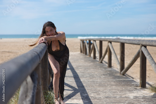 Young, beautiful woman with long brown hair, wearing a swimming costume and a black silk sarong, rests her outstretched arm on the railing of the walkway leading to the beach. Sea in the background.