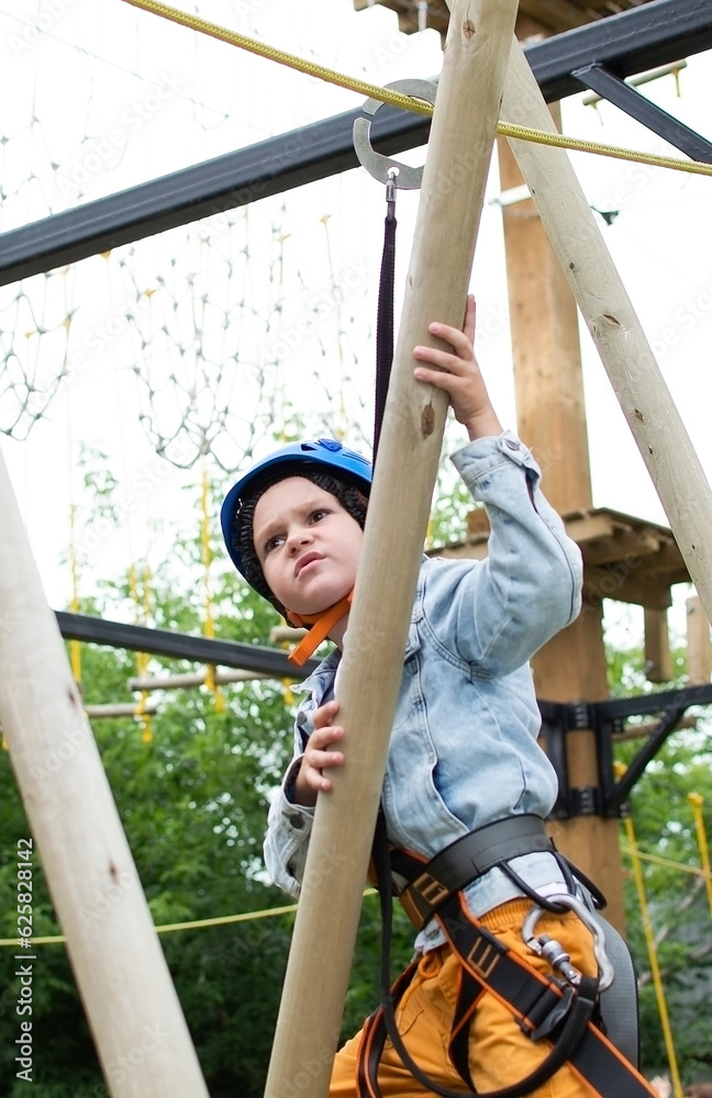 Fototapeta premium Child in forest adventure park. An outdoor entertainment center for children engaged in agility and rock climbing. Children enjoy spending time outdoors on warm sunny summer day. Vertical photography.