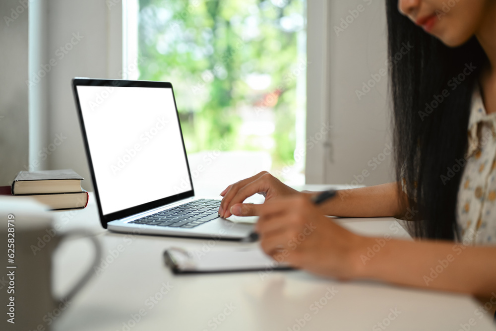 Side view of female freelancer working online on laptop and writing important notes Side view of female freelancer working online on laptop and writing important notes