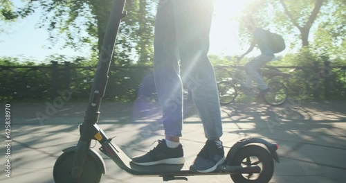 Woman on electric scooter overtakes man on bicycle. City park, embankment, summer weather, bright sun.