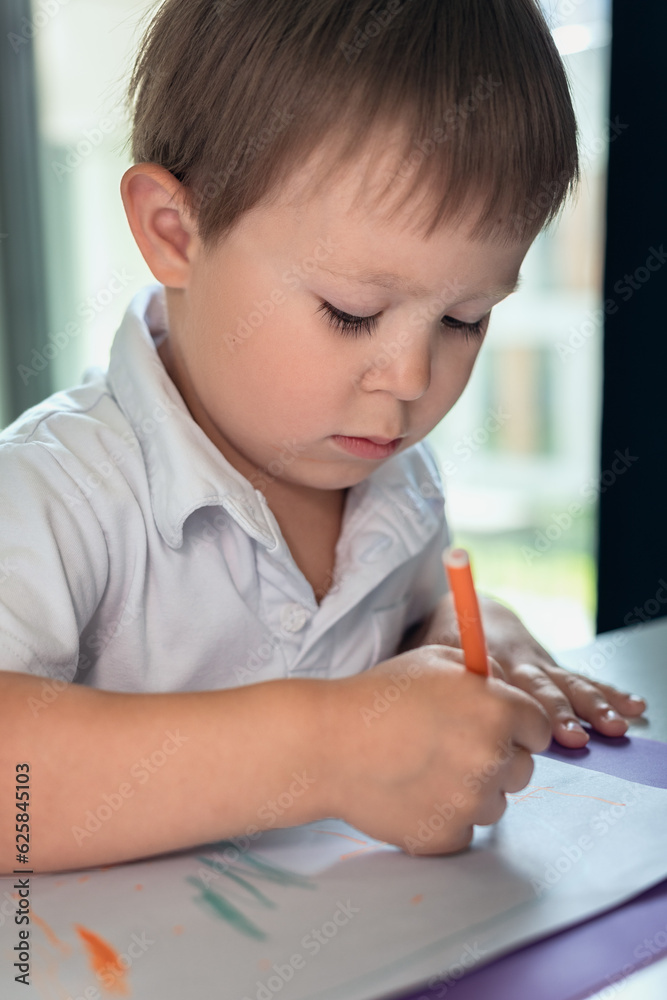 a small preschool child sits at a desk and draws on a white sheet of paper a small preschool child sits at a desk and draws on a white sheet of paper