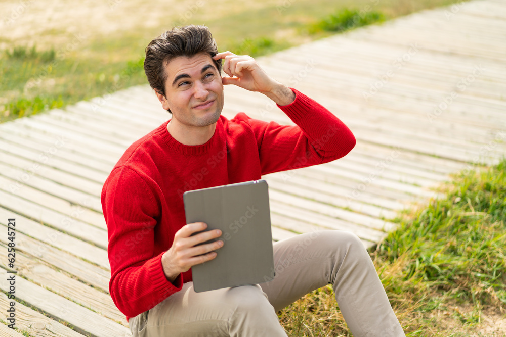 Fototapeta premium Young handsome man holding a tablet at outdoors having doubts and with confuse face expression