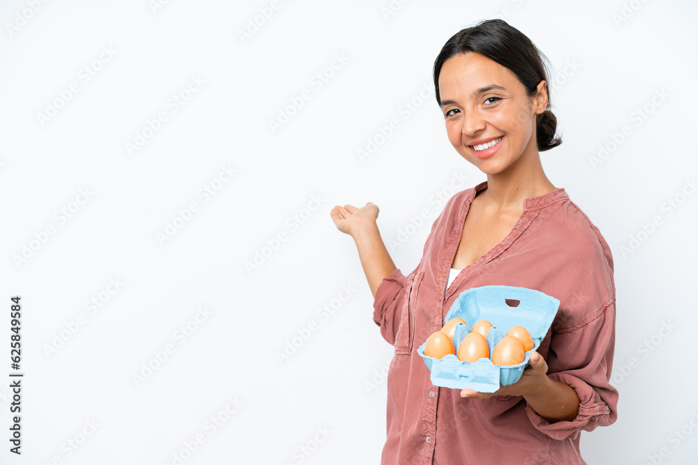 Young hispanic woman holding eggs isolated on white background extending hands to the side for inviting to come Young hispanic woman holding eggs isolated on white background extending hands to the side for inviting to come