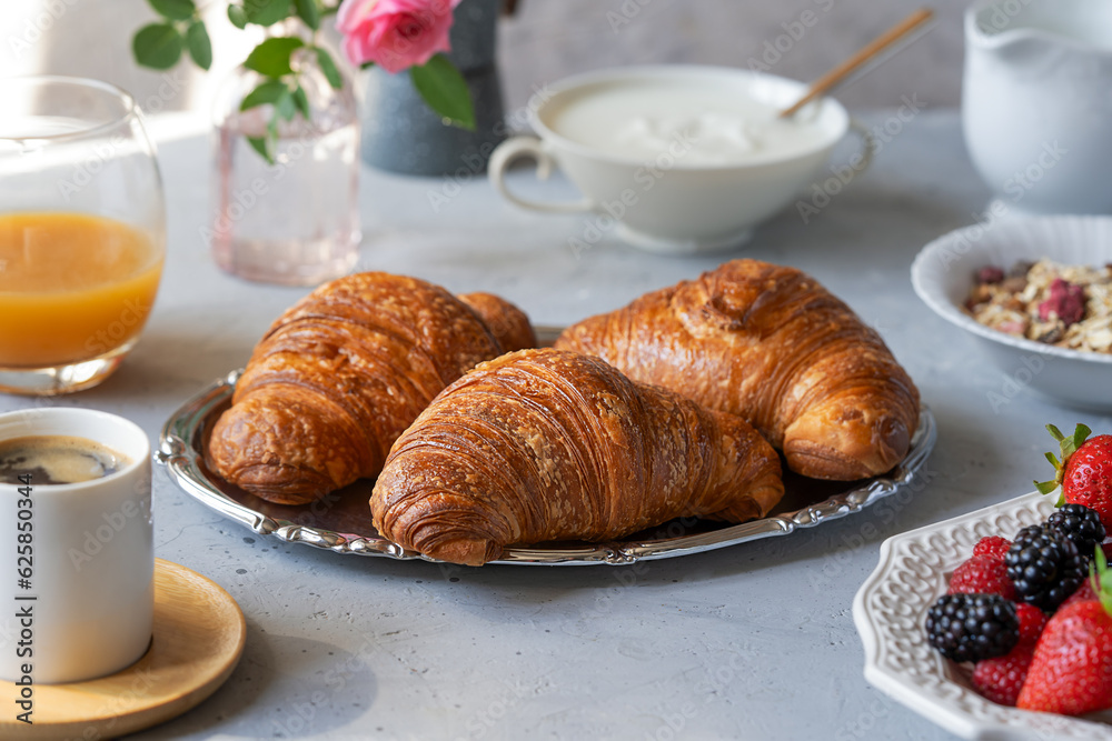 Breakfast in the morning with fresh baked croissants, berries, muesli, coffee, milk, yogurt, honey and juice over grey background Breakfast in the morning with fresh baked croissants, berries, muesli, coffee, milk, yogurt, honey and juice over grey background