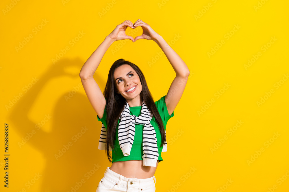 Photo of dreamy shiny girl dressed green t-shirt rising arms showing heart looking empty space isolated yellow color background