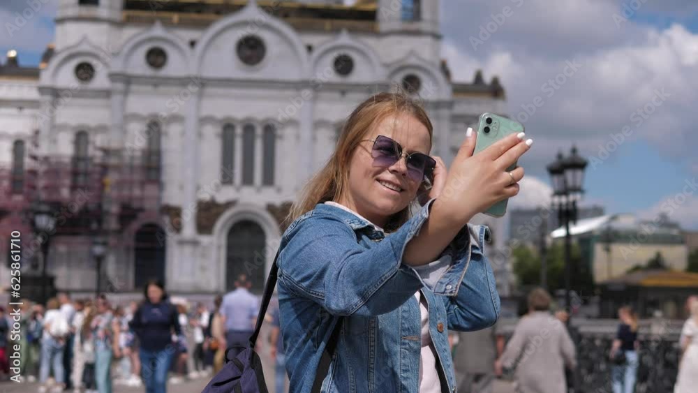 Portrait of a young woman who shoots a video standing against the background of a church and a lot of people. A tourist takes a selfie on her phone with the city during a trip. Portrait of a young woman who shoots a video standing against the background of a church and a lot of people. A tourist takes a selfie on her phone with the city during a trip.
