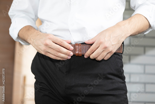 Young businessman wearing pant belt in the room, close-up photo. Preparation for new busy day