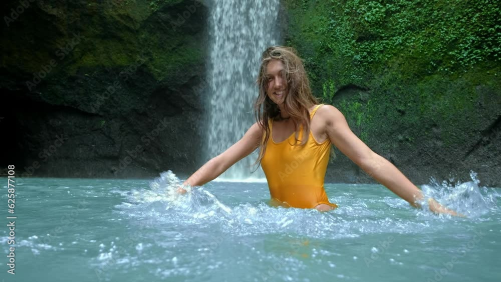 Happy young women makes splashes at waterfall background. Behind the girl, she smiles and plays with splashes of water, relaxing and enjoying the waterfalls of tropical nature. The concept of a Happy young women makes splashes at waterfall background. Behind the girl, she smiles and plays with splashes of water, relaxing and enjoying the waterfalls of tropical nature. The concept of a