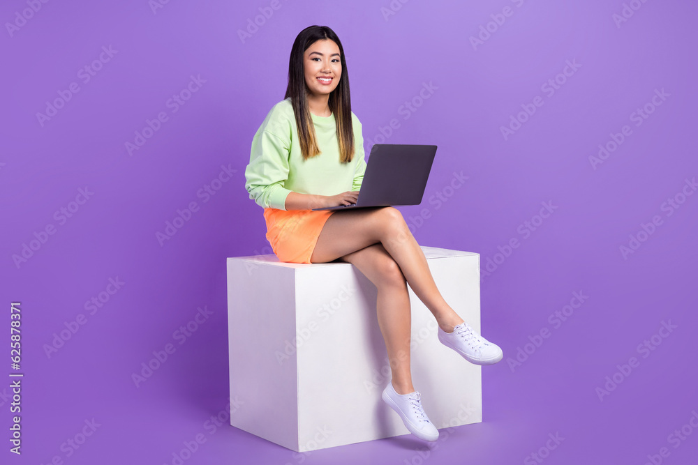 Full length photo of optimistic adorable smart girl wear stylish pullover sit on platform with laptop isolated on violet color background Full length photo of optimistic adorable smart girl wear stylish pullover sit on platform with laptop isolated on violet color background
