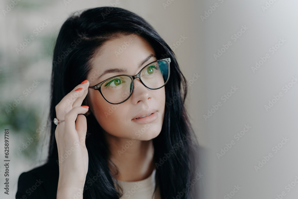 Serious brunette with spectacles, focused on screen, browsing web, creating projects indoors, office setting.
