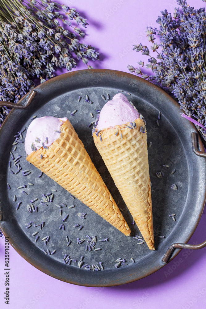 Two lavender ice creams in waffle cones on a tin tray and bouquets of dry lavender on a lilac background.