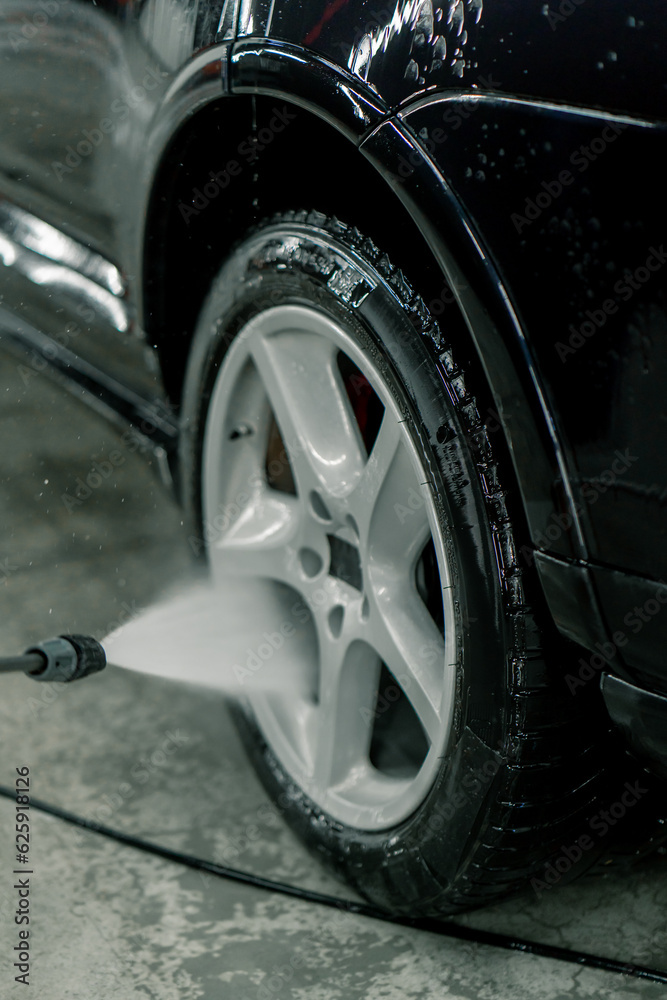 Close-up of a male car wash employee washing the rim and tire of a black luxury car using a high-pressure washer 