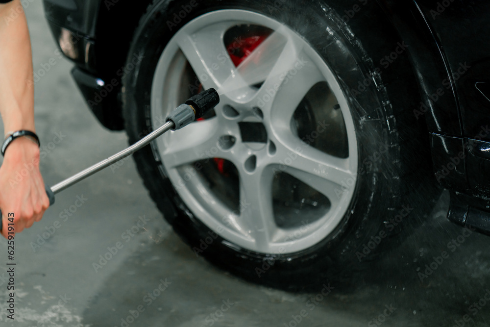 Close-up of a male car wash employee washing the rim and tire of a black luxury car using a high-pressure washer 