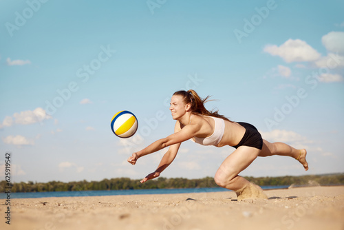 Competitive young woman athlete playing beach volleyball, hitting ball with hands and falling down on sand. Match. Concept of sport, active and healthy lifestyle, hobby, summertime, ad