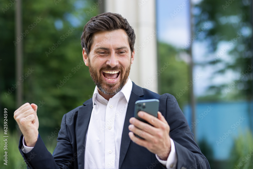 Mature experienced businessman with phone celebrating success victory triumph, boss in business suit received good notification online, man outside office building holding hand up winner gesture Mature experienced businessman with phone celebrating success victory triumph, boss in business suit received good notification online, man outside office building holding hand up winner gesture