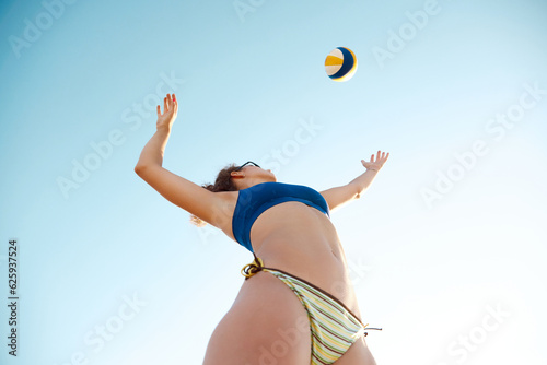 Dynamic bottom view image of young sportive woman playing beach volleyball, hitting ball in a jump over sky background. Concept of sport, active and healthy lifestyle, hobby, summertime, ad