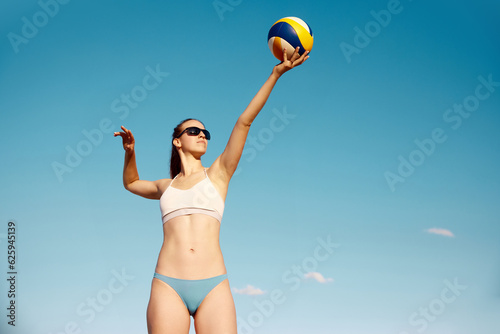 Bottom view dynamic image of young woman in swimsuit serving ball, play in g beach volleyball over blue sky background. Concept of sport, active and healthy lifestyle, hobby, summertime, ad #625945139