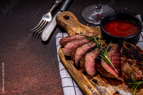 Grilled meat steak on dark table, Pork, veal, lamb, beef steak medium, medium well on cutting board, with spices, ketchup bbq sauce, wine glass. Dinner in steak restaurant menu, top view copy space