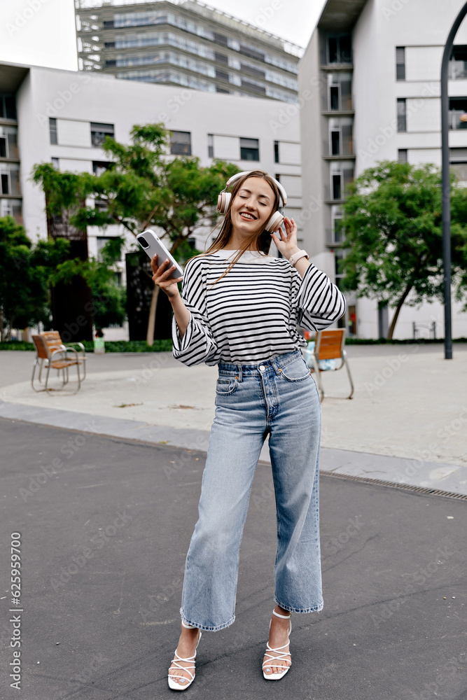Full-length photo of stylish good-looking girl in jeans and summer shirt on heels is holding smartphone and enjoying music in headphones on the street in city