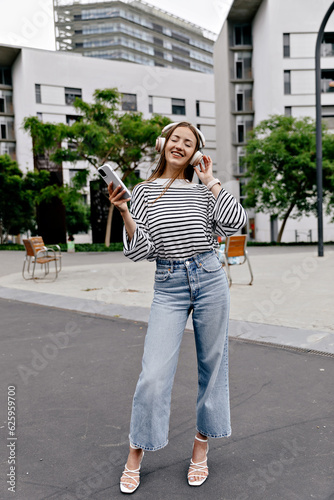 Full-length photo of stylish good-looking girl in jeans and summer shirt on heels is holding smartphone and enjoying music in headphones on the street in city © PhotoBook