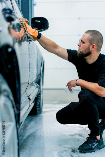A car wash worker uses a microfiber cloth to wash the front headlight of a black luxury car with car wash shampoo