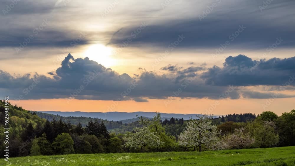 Sunny sunset in a cloudy sky illuminates a hilly valley with blooming orchards, meadows and forests Sunny sunset in a cloudy sky illuminates a hilly valley with blooming orchards, meadows and forests