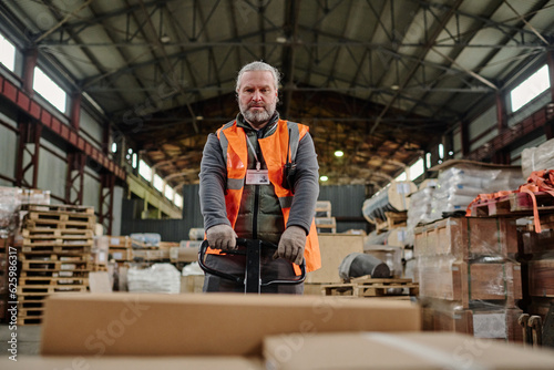 Mature warehouse worker carrying containers on loader during his work in warehouse