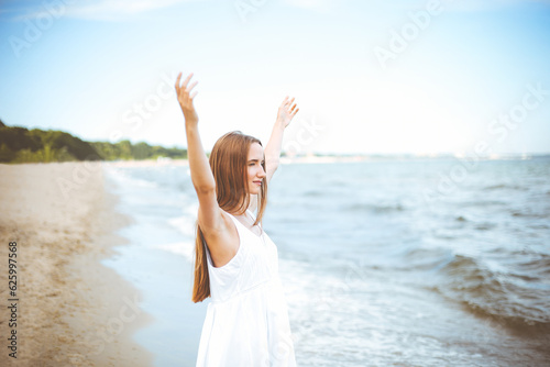 Happy smiling woman in free happiness bliss on ocean beach standing with raising hands. Portrait of a multicultural female model in white summer dress enjoying nature during travel holidays vacation