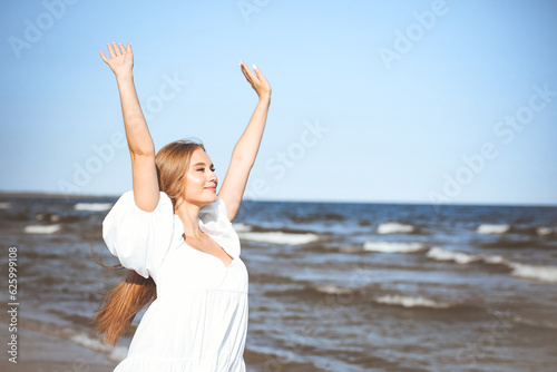 Happy smiling beautiful woman on the ocean beach standing in a white summer dress, raising hands