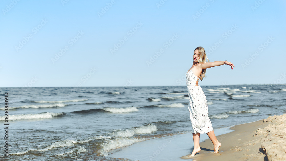 Happy blonde beautiful woman on the ocean beach standing in a white summer dress, open arms