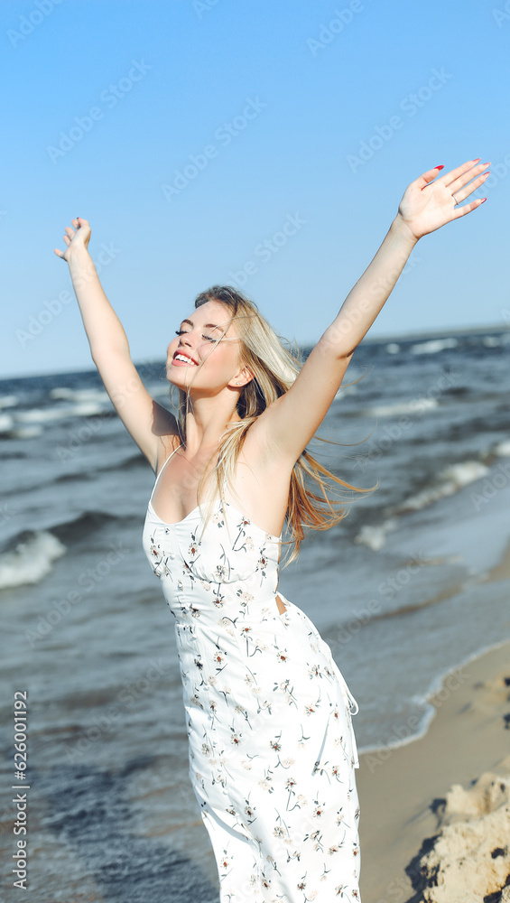 Happy blonde beautiful woman on the ocean beach standing in a white summer dress, raising hands Happy blonde beautiful woman on the ocean beach standing in a white summer dress, raising hands