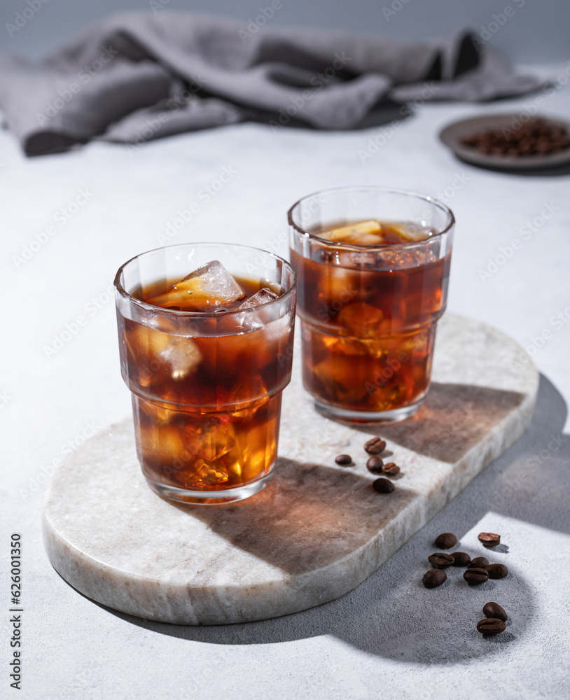 Cold brew coffee in a two glasses with ice on a marble board on a light background with coffee beans, shadows and napkin. Concept summer craft refreshing homemade drink