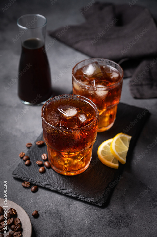 Cold brew coffee in a two glasses with ice and lemon on a dark concrete background with coffee beans, bottle and napkin. Concept summer craft refreshing homemade drink Cold brew coffee in a two glasses with ice and lemon on a dark concrete background with coffee beans, bottle and napkin. Concept summer craft refreshing homemade drink