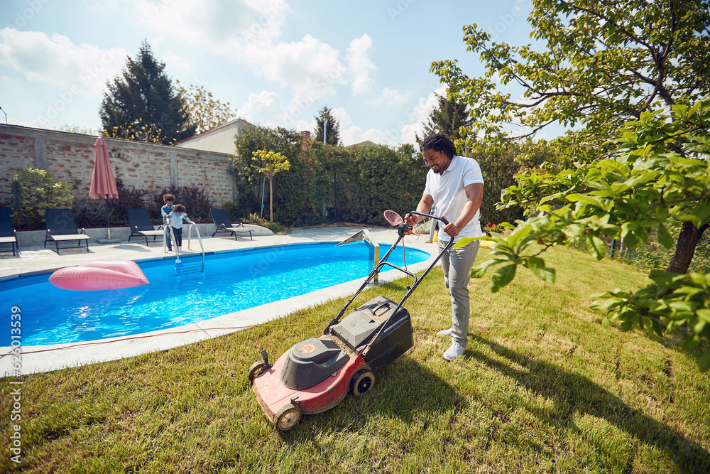man cutting grass with electric grass mower by the pool, with his kids