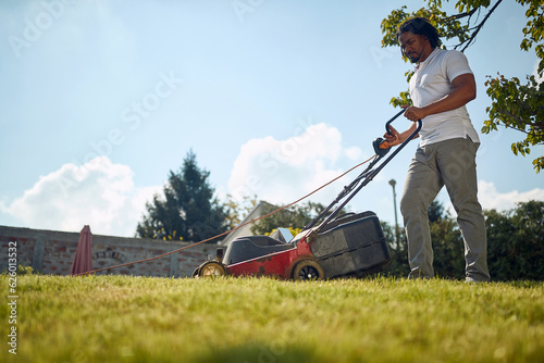 Handsome young african american man mowing grass outdoors, making the backyard beutiful and neat.