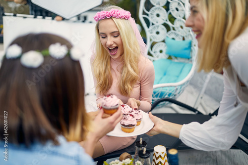 Young joyful women on a bachelorette party enjoying cupcakes with frosting, sitting outdoors on a sunny day.