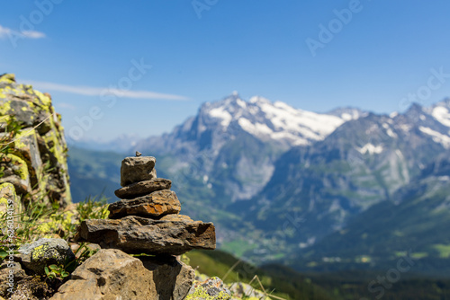 A small rock cairn balances on a boulder with a view of mountains of the Swiss alps and blue skies in the background © Harrison
