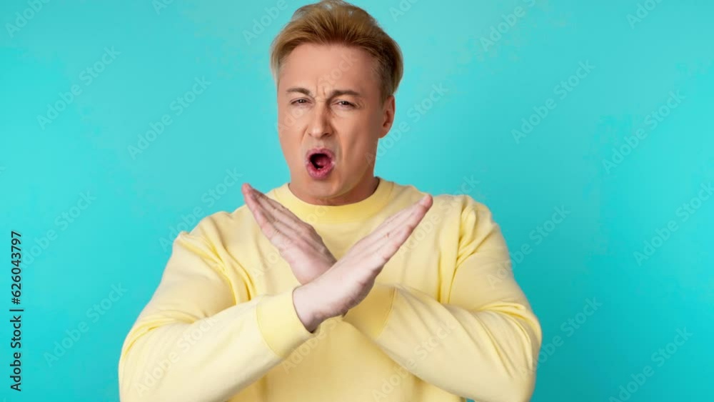 Displeased man says no in stop gesture crossing hands. Guy doing negative sign nodding no, angry face, rejection expression in yellow sweatshirt. Indoor studio shot isolated on blue background. Displeased man says no in stop gesture crossing hands. Guy doing negative sign nodding no, angry face, rejection expression in yellow sweatshirt. Indoor studio shot isolated on blue background.