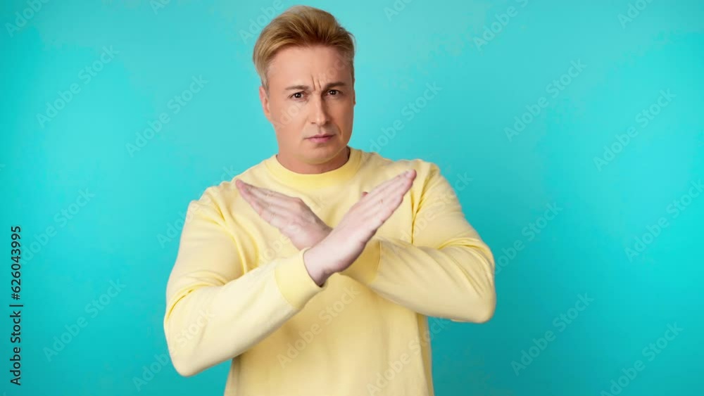 Displeased man says no in stop gesture crossing hands. Guy doing negative sign nodding no, angry face, rejection expression in yellow sweatshirt. Indoor studio shot isolated on blue background. Displeased man says no in stop gesture crossing hands. Guy doing negative sign nodding no, angry face, rejection expression in yellow sweatshirt. Indoor studio shot isolated on blue background.