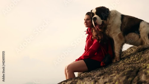Cute shot of pretty woman with a dog saint bernard sitting on the peak and enjoying beautiful mountains view and cloudy sky together Animals and human friendship. High quality 4k footage photo