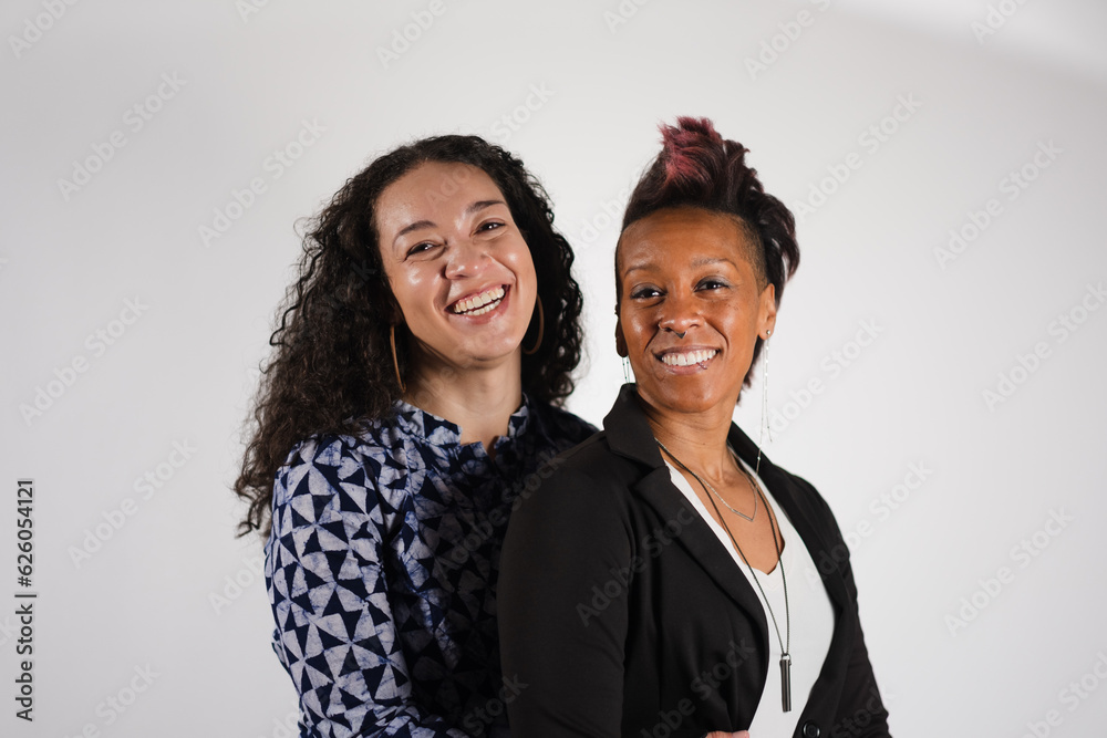 Standing together against a white studio backdrop, a multiracial, lesbian, and queer couple poses for engagement photos. One women has her arms in an embrace around her partner's waist. Standing together against a white studio backdrop, a multiracial, lesbian, and queer couple poses for engagement photos. One women has her arms in an embrace around her partner's waist.