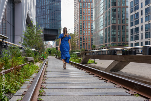 A woman in blue maxi dress walking on an old railing turned into a pathway - the famous High Line in New York. The railing is overgrown with greenery. Some benches on the sides. Rearranging the city.