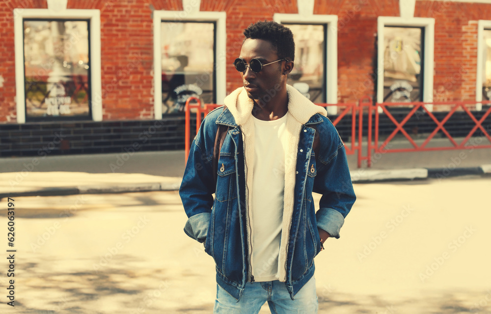 Portrait of stylish young african man posing wearing denim jacket, backpack on city street background Portrait of stylish young african man posing wearing denim jacket, backpack on city street background