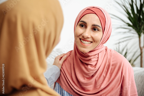 Portrait of a young beautiful Muslim woman who is sitting on a sofa in her house has a pleasant conversation with her Islamic friend. The girl takes the hugs of meeting and held a conversation