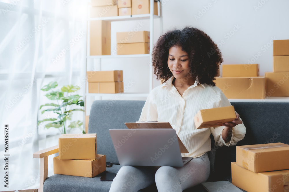 Smiling young business owner woman prepare parcel box and check online orders of product for deliver to customer on laptop computer at home. Shopping Online concept. Startup small business SME.